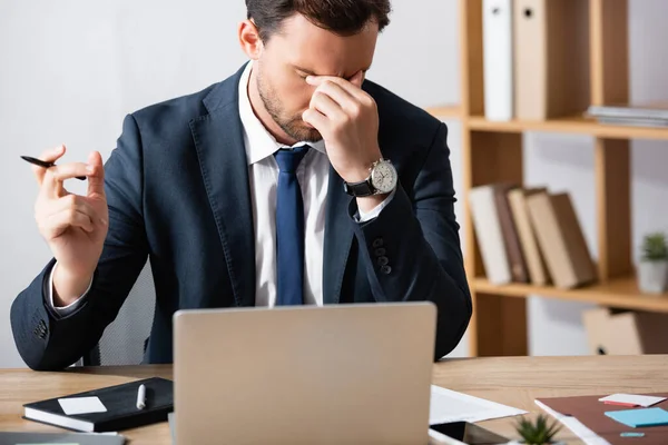 Hombre de negocios cansado con migraña, cogido de la mano cerca de los ojos, mientras está sentado en el lugar de trabajo sobre un fondo borroso - foto de stock