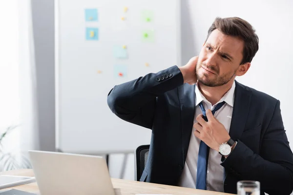 Бизнесмен с болью в шее развязывает галстук, сидя на рабочем месте с размытым флипчартом на заднем плане — стоковое фото