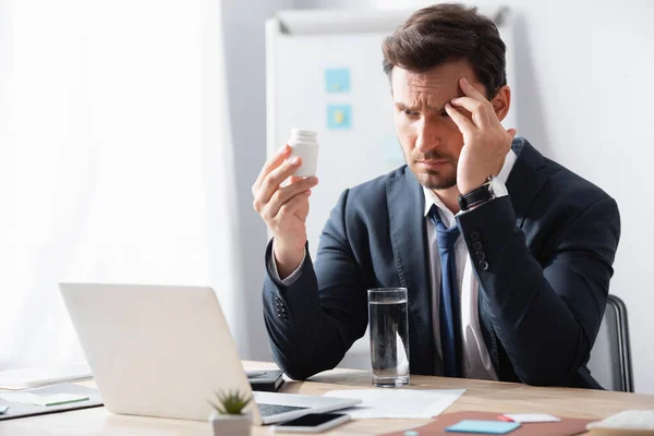 Müder Geschäftsmann mit Kopfschmerzen beim Betrachten von Medikamenten, während er am Arbeitsplatz mit verschwommenem Flipchart im Hintergrund sitzt — Stockfoto
