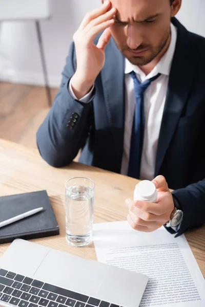 Businessman with migraine looking at medication, while sitting at workplace on blurred background — Stock Photo