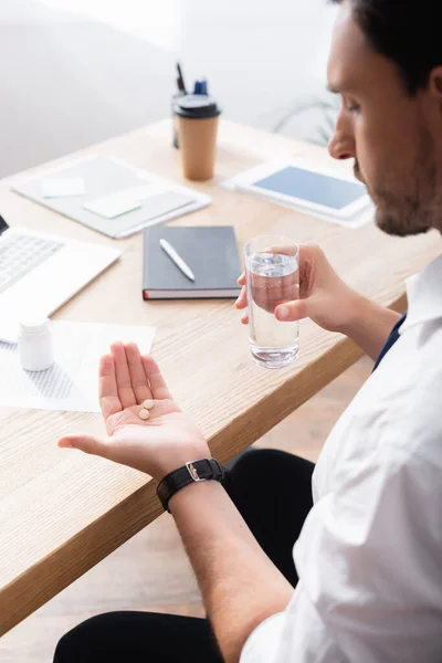 Homme d'affaires tenant un verre d'eau et regardant des pilules sur la paume de la main, assis sur le lieu de travail sur un fond flou — Photo de stock