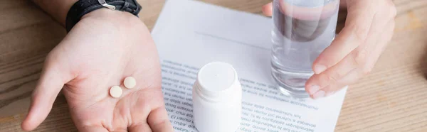 Vista recortada del hombre sosteniendo un vaso de agua y pastillas cerca del contrato comercial sobre fondo de madera, pancarta - foto de stock