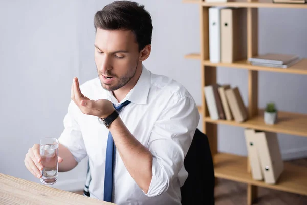 Businessman with glass of water, taking medicines, while sitting at table in office on blurred background — Stock Photo