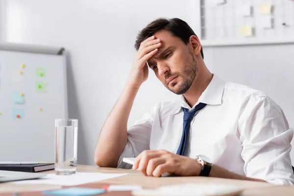 Geschäftsmann mit Kopfschmerzen hält Smartphone in der Hand, sitzt am Arbeitsplatz mit verschwommenem Schreibtisch im Vordergrund — Stockfoto