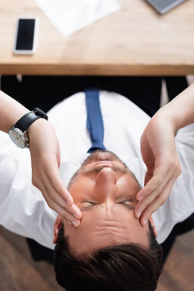 Vista superior del hombre de negocios con dolor de cabeza sentado en el lugar de trabajo sobre fondo borroso - foto de stock