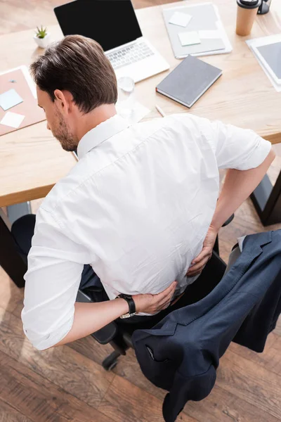 Overhead view of businessman with hands on painful lower back sitting at workplace in office — Stock Photo