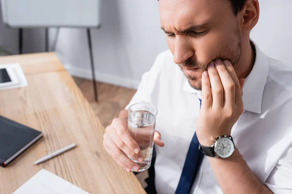 Businessman holding glass of water and suffering from toothache, while sitting at workplace on blurred background — Stock Photo