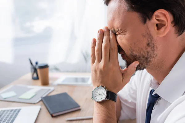 Vista lateral del hombre de negocios cansado con las manos en oración, que sufre de dolor de cabeza con el lugar de trabajo borroso en el fondo - foto de stock