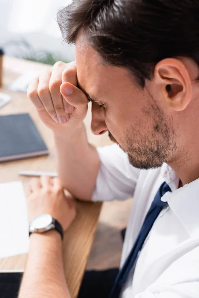 Businessman with closed eyes leaning on hand, suffering from headache, while sitting at workplace on blurred background — Stock Photo
