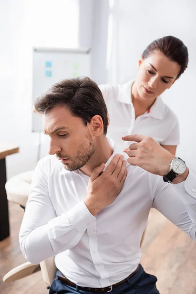 Masajista mirando hacia atrás del cliente señalando con el dedo el doloroso cuello, mientras está sentado en una silla de masaje sobre un fondo borroso - foto de stock