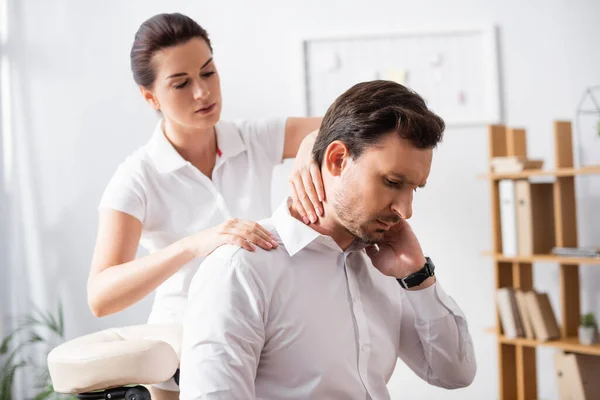 Female massage therapist massaging hurting neck of businessman sitting on massage chair on blurred background — Stock Photo