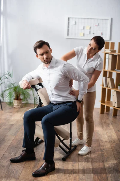 Masajista enfocado masajeando lastimando la espalda del hombre de negocios con la mano en la cadera, sentado en silla de masaje en la oficina - foto de stock