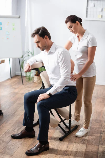 Female massage therapist massaging painful back of businessman sitting on massage chair in office — Stock Photo