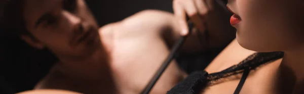 Donna sexy con labbra rosse vicino al fidanzato con frusta bdsm su sfondo sfocato isolato su nero, banner — Foto stock