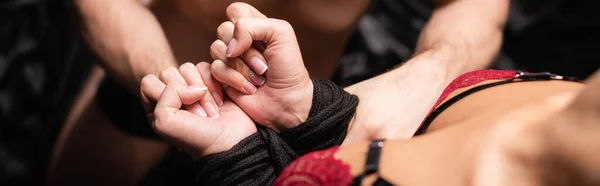 Cropped view of man tying hands of girlfriend in bra with rope on blurred background, banner — Stock Photo