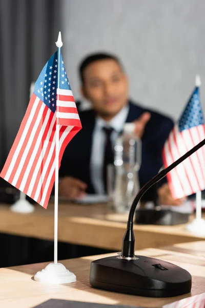 Bandeira americana pequena com microfone na mesa com homem indiano borrado no fundo — Fotografia de Stock