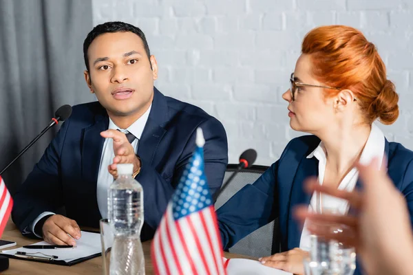 Indian politician gesturing, while talking with colleague, sitting near redhead woman in boardroom on blurred foreground — Stock Photo