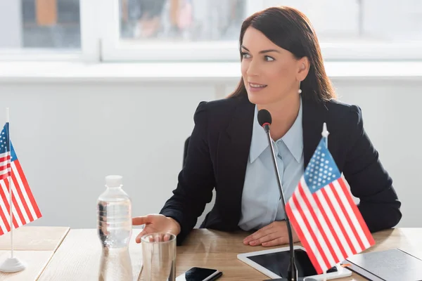 Lächelnde Frau in offizieller Kleidung, die wegschaut, während sie in Mikrofon spricht, neben amerikanischen Flaggen im Sitzungssaal sitzt — Stockfoto