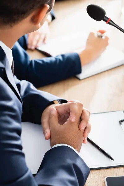 Ausgeschnittene Ansicht eines indischen Politikers mit geballten Händen auf Klemmbrett mit leerem Blatt Papier, das neben Mikrofon auf verschwommenem Hintergrund sitzt — Stockfoto