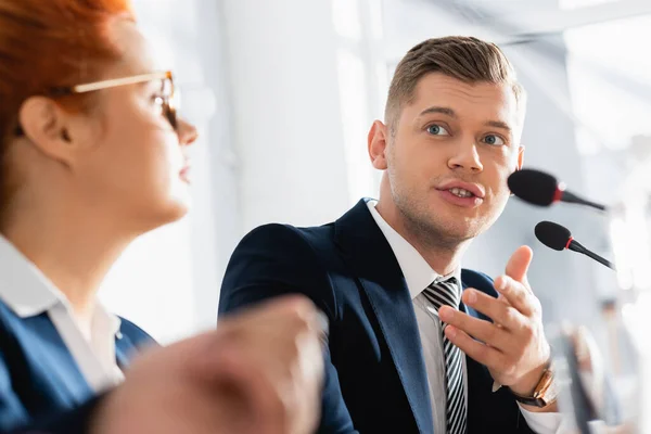 Politician gesturing, while talking with colleague, sitting near microphone with blurred woman on foreground — Stock Photo