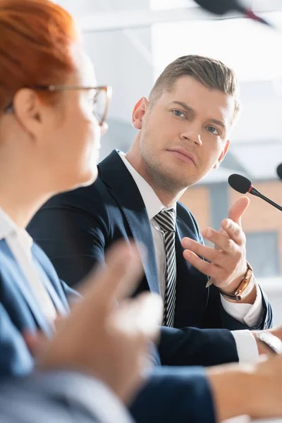 Serious politician gesturing, while looking at colleague, sitting near microphone with blurred woman on foreground — Stock Photo