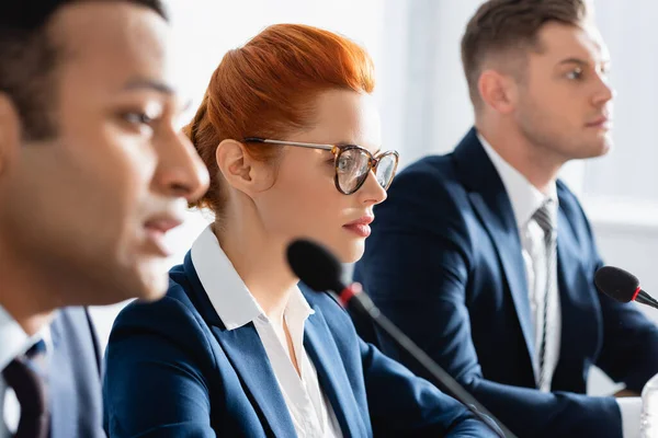 Femme politique rousse aux lunettes, assise près d'un homme indien parlant au micro au premier plan flou — Photo de stock