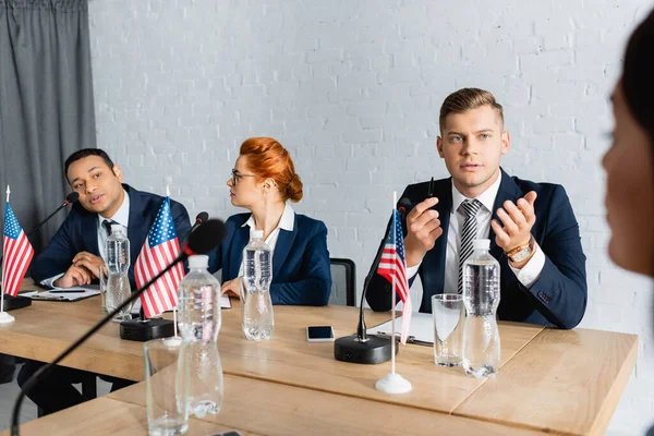 Político indio mirando a una colega, mientras habla en micrófono, sentada cerca de miembros del partido político en un primer plano borroso - foto de stock