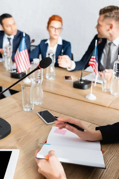 Female politician with pen and notebook sitting near smartphone and microphone with blurred colleagues working on background — Stock Photo