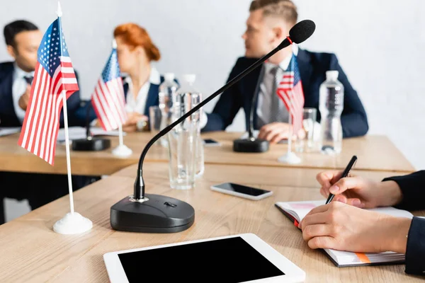 Politiker schreibt in Notizbuch, während er neben Mikrofon und digitalem Tablet sitzt und verschwommene Menschen im Hintergrund reden — Stockfoto