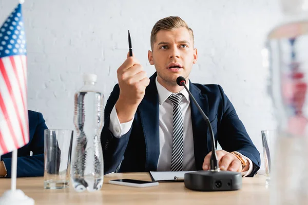 Serious politician gesturing with pen, while speaking in microphone and sitting near colleague on blurred foreground — Stock Photo