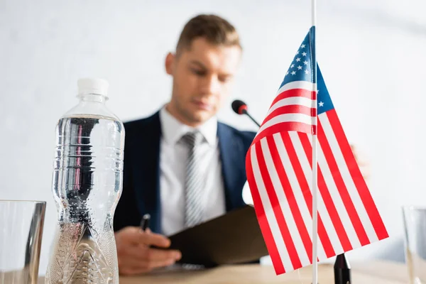 Garrafa de água e pequena bandeira americana na mesa com político desfocado no fundo — Fotografia de Stock