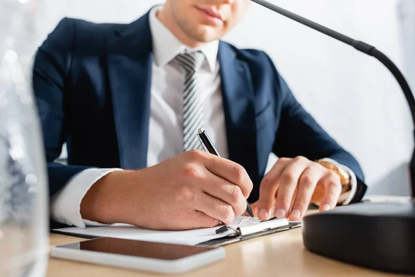 Ausgeschnittene Ansicht eines Politikers, der auf Klemmbrett schreibt, während er mit Mikrofon und Handy auf verschwommenem Vordergrund am Tisch sitzt — Stockfoto