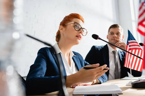 Político reflexivo mirando a una colega hablando en micrófono, mientras está sentada en la sala de juntas en un primer plano borroso — Stock Photo