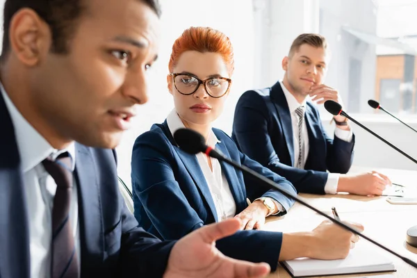 Rosse politico femminile guardando collega indiana che parla al microfono durante la riunione del partito politico nella sala del consiglio di amministrazione in primo piano sfocato — Stock Photo