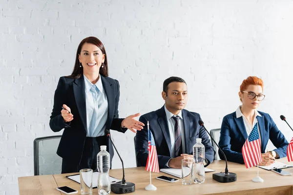Smiling female politician with open arms talking, while standing near interracial colleagues in boardroom — Stock Photo