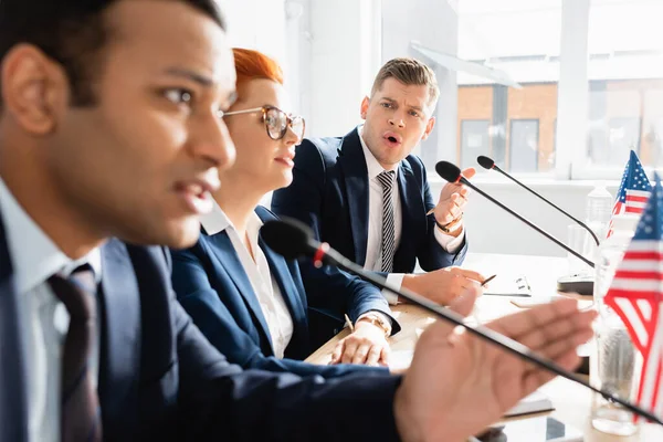 Político irritado com boca aberta olhando para colega falando em microfone, enquanto sentado à mesa na sala de reuniões em primeiro plano desfocado — Fotografia de Stock