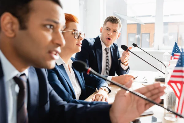 Politico irritato a bocca aperta che guarda il collega che parla al microfono, seduto a tavola in sala riunioni in primo piano sfocato — Foto stock