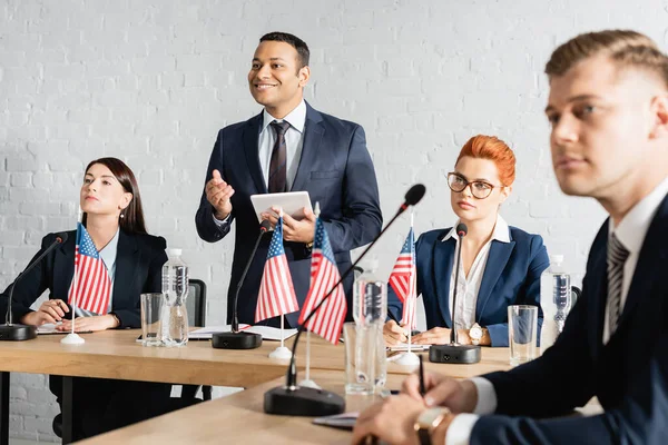 Político indio feliz mirando hacia otro lado, mientras está de pie con la tableta digital cerca de colegas en la sala de juntas - foto de stock