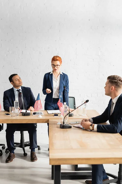 Politiker blicken auf rothaarige Kollegin, die mit Stift gestikuliert, während sie während einer Parteiversammlung im Sitzungssaal spricht — Stockfoto