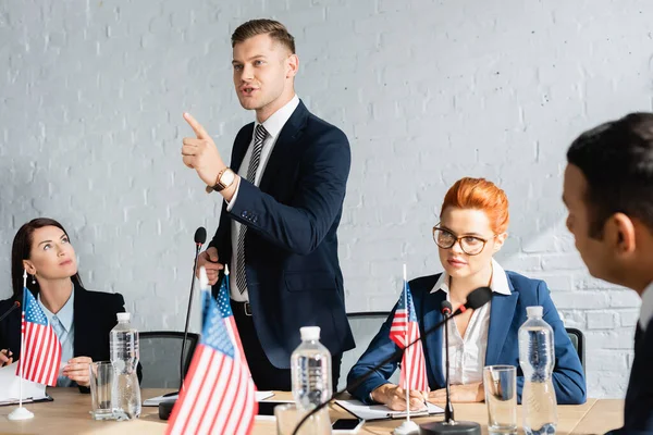 Lächelnder Politiker, der mit dem Finger zeigt, spricht, während er neben einem Kollegen steht, der auf Klemmbrett im verschwommenen Vordergrund schreibt — Stockfoto