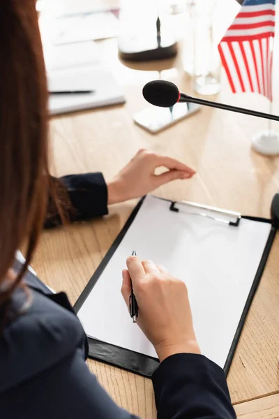 Vue recadrée de la femme brune tenant un stylo assis près du presse-papiers avec du papier blanc et un microphone sur fond flou — Photo de stock