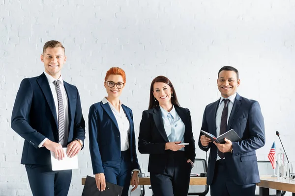 Lächelnde multikulturelle Politiker, die in die Kamera schauen, während sie im Sitzungssaal andere Sachen in der Hand halten — Stockfoto