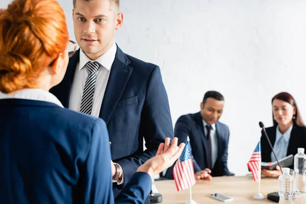 Woman gesturing while talking to colleagues near multicultural party members on blurred background — Stock Photo