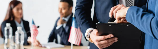 Politiker zeigt mit dem Finger auf Klemmbrett in der Nähe von Kollegen, die auf verschwommenem Hintergrund reden, Banner — Stockfoto