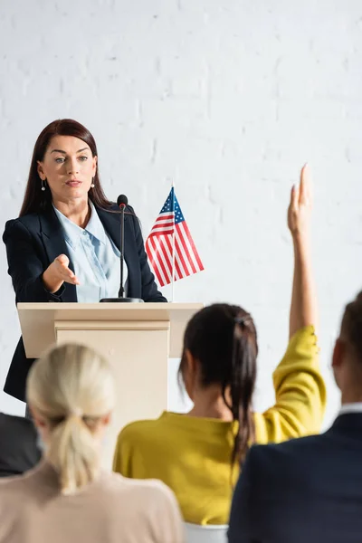 Speaker pointing with hand at woman with raised hand in conference hall, blurred foreground — Stock Photo