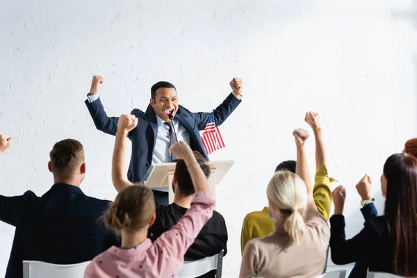 Excited indian speaker showing winner gesture while standing in front of voters in conference hall, blurred foreground — Stock Photo