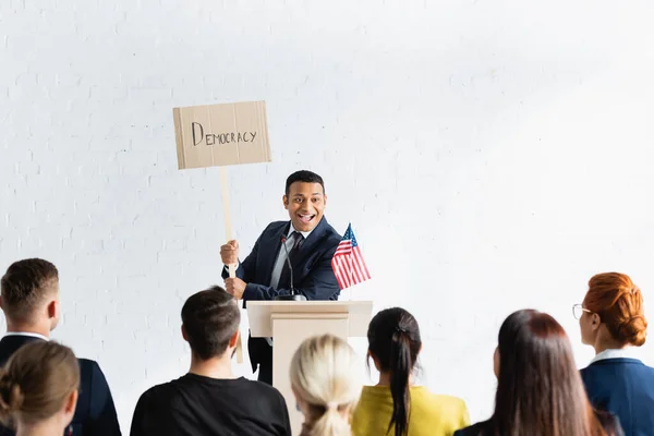 Orador indio emocionado sosteniendo pancarta con la inscripción de la democracia delante de los votantes en la sala de conferencias, borrosa primer plano — Stock Photo