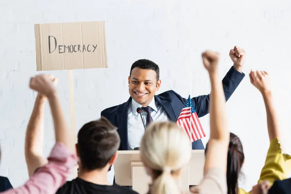 Orador indio emocionado sosteniendo pancarta con la inscripción de la democracia y mostrando el gesto ganador frente a los votantes, borrosa primer plano - foto de stock