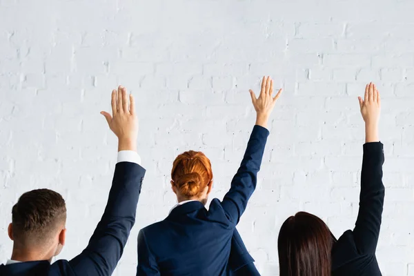 Vue arrière des femmes et des hommes votant avec les mains dans l'air contre le mur de briques blanches — Photo de stock