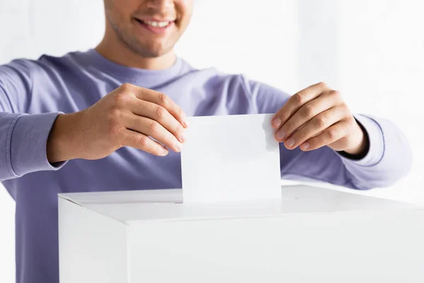 Cropped view of smiling man inserting ballot into polling box on blurred background — Stock Photo